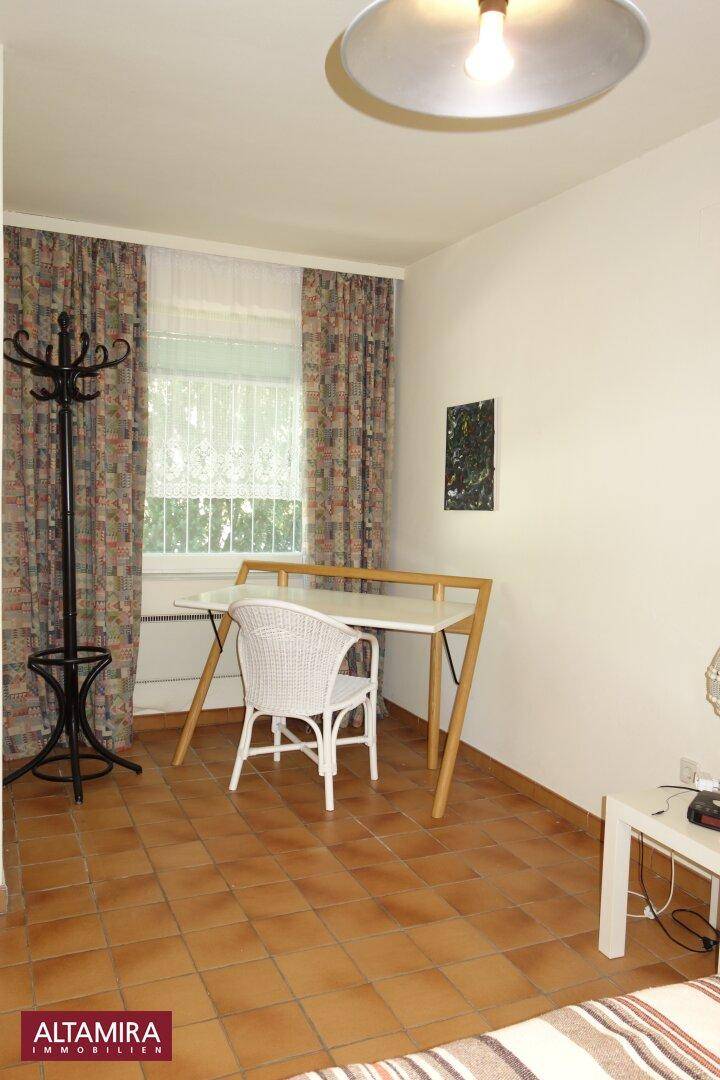 Helles Schlafzimmer mit Terrakottafliesen, Schreibtisch und Fenster mit Vorhängen.