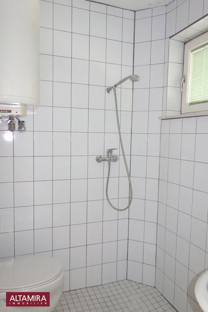 Funktionales Badezimmer mit Dusche, WC und Fenster, komplett gefliest in Weiß.