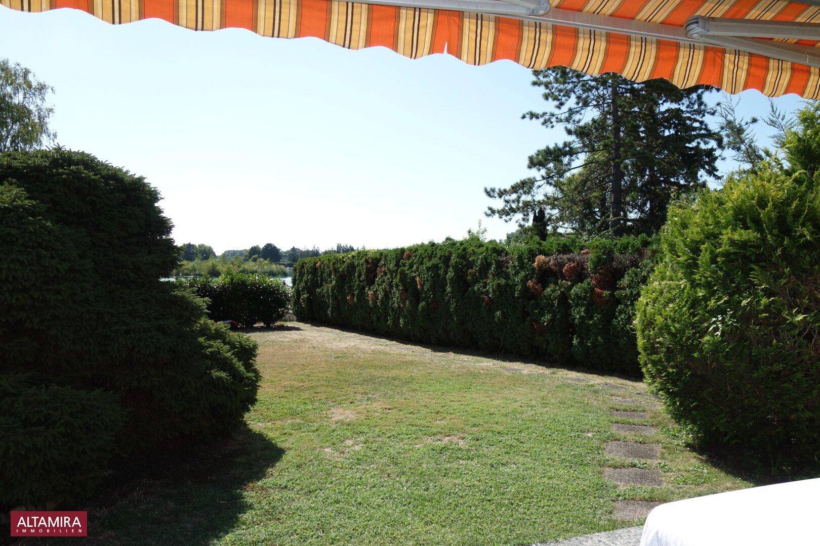 Gepflegter Garten mit hoher Hecke und weitem Blick auf den See im Hintergrund.