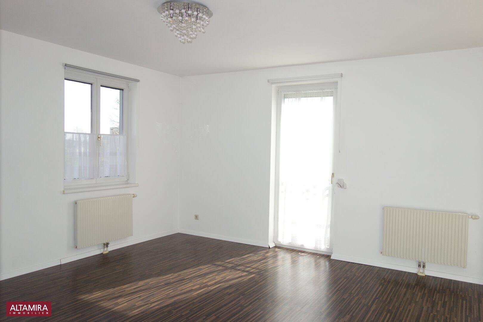 Geräumiges Zimmer mit dunklem Laminatboden, großem Fenster und Zugang zu einem Außenbereich.
