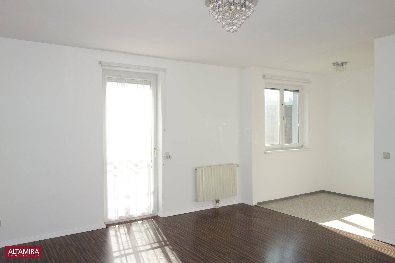 Heller Wohnbereich mit dunklem Laminatboden und einem Übergang zu einem weiteren Zimmer.