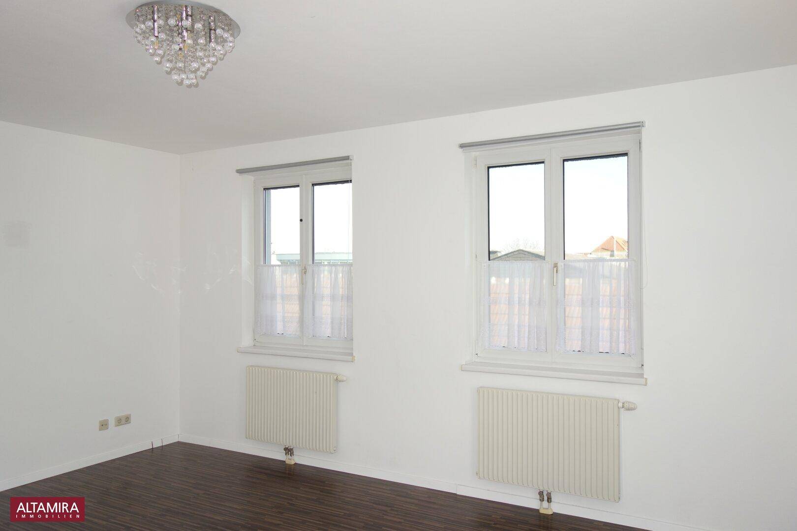 Helles Zimmer mit zwei Fenstern, dunklem Laminatboden und einem Kronleuchter an der Decke.