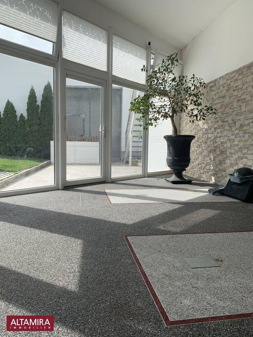 Heller Innenraum mit großen Glasfronten, einem dekorativen Baum und gemusterter Bodenfliese.
