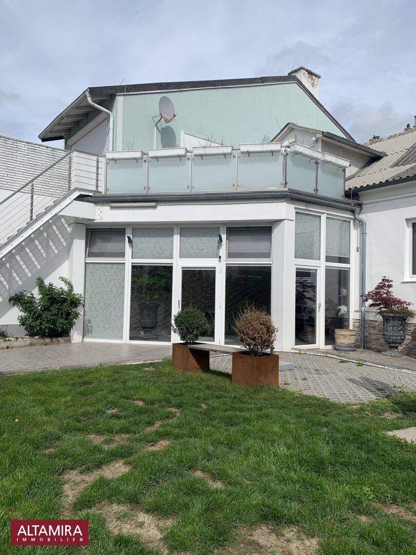 Außenansicht des Gebäudes mit einer Terrasse im Obergeschoss und einem grünen Gartenbereich.