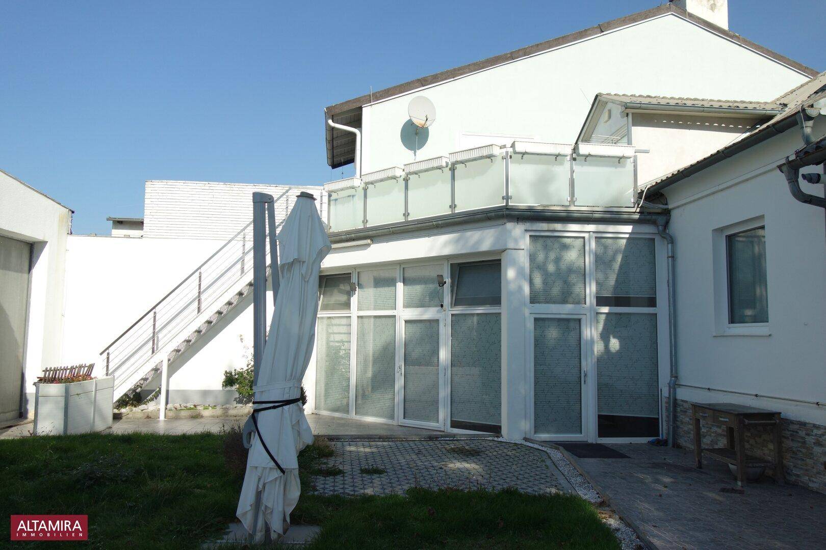 Außenansicht des Gebäudes mit einer Terrasse im Obergeschoss und einem ungepflegten Gartenbereich.