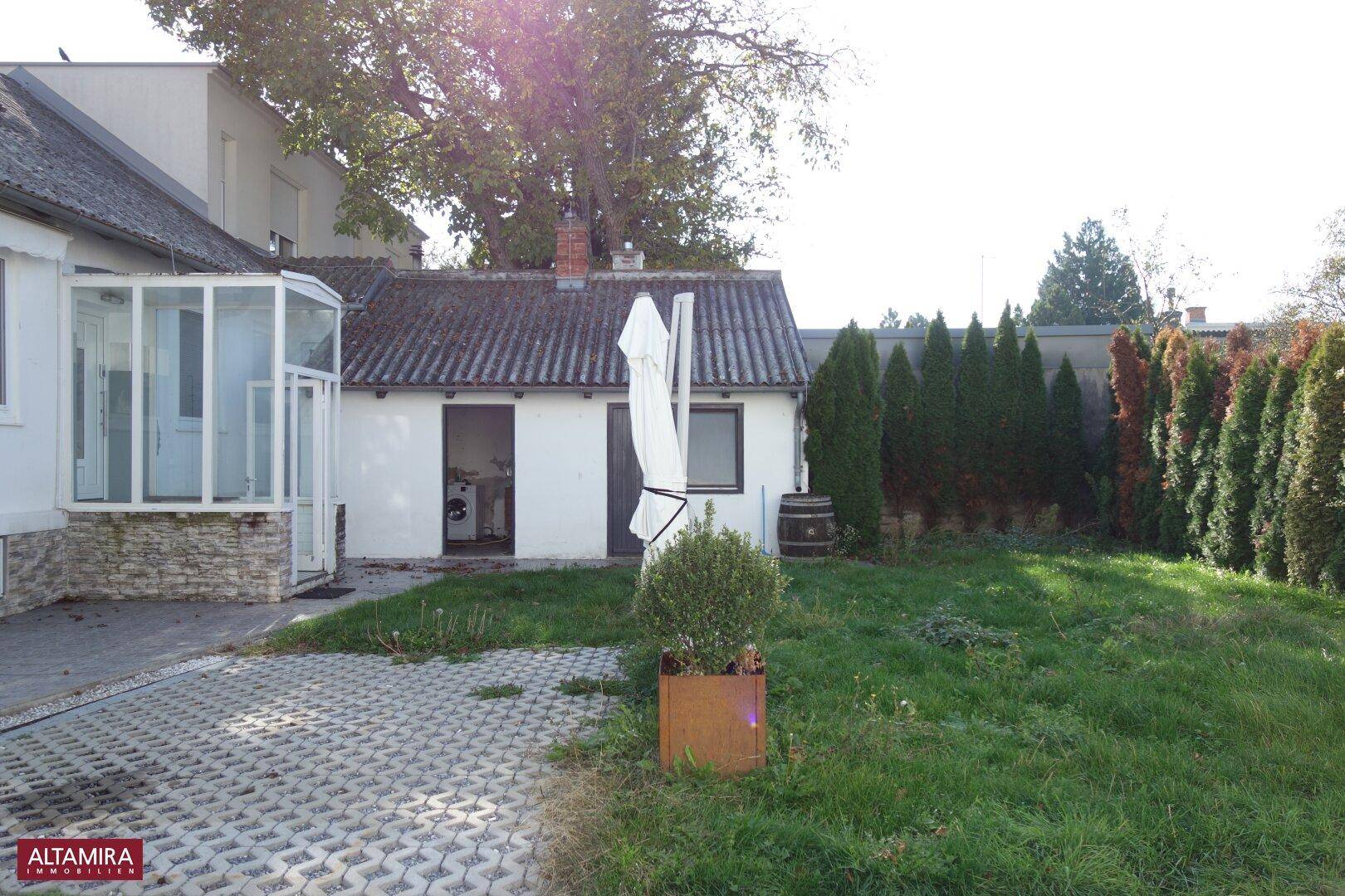 Gartenbereich mit Rasenfläche, hohen Hecken und einem kleinen Nebengebäude im Hintergrund.