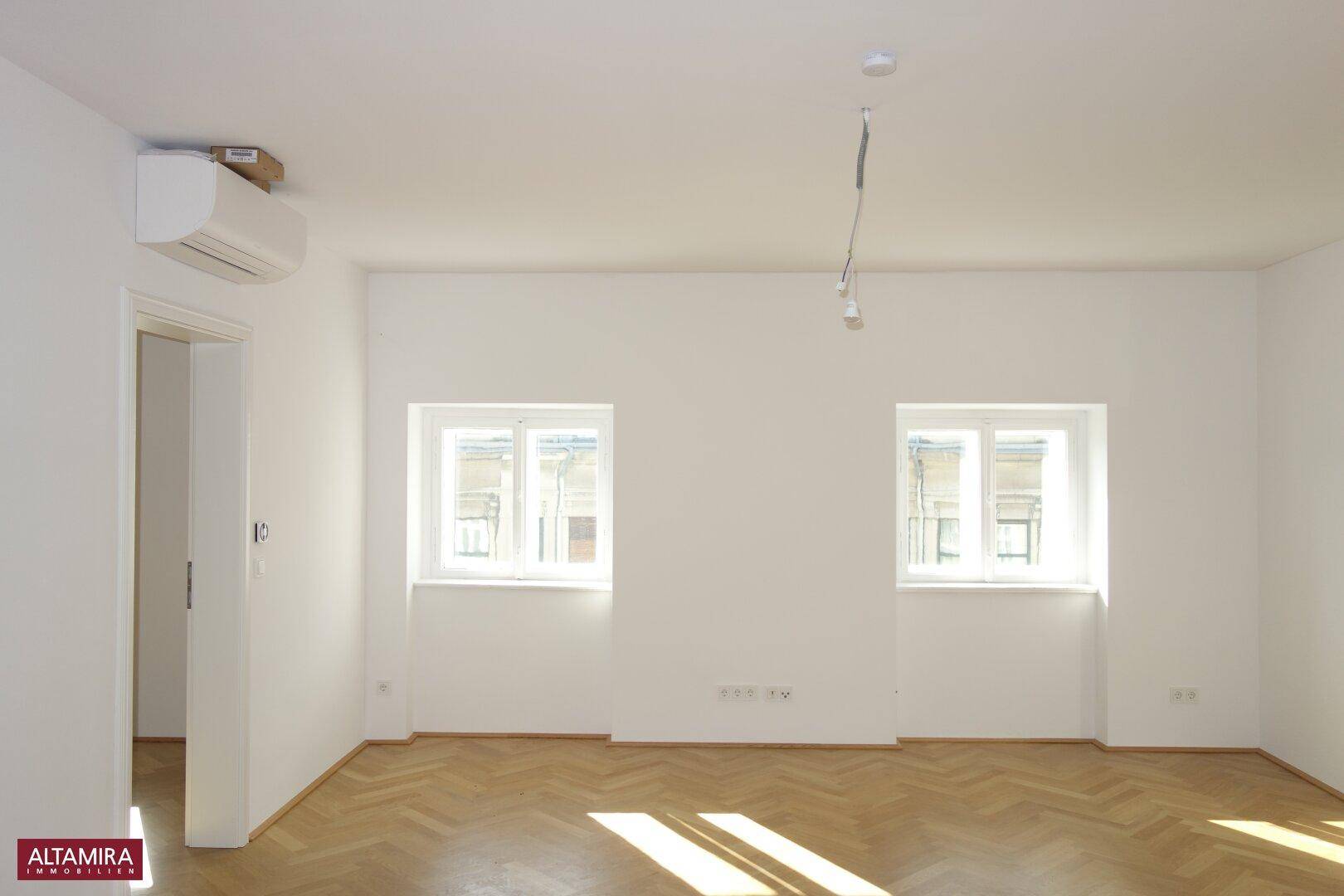 Geräumiger, heller Raum mit Fischgrätparkett, Klimaanlage und zwei Fenstern für viel Tageslicht.