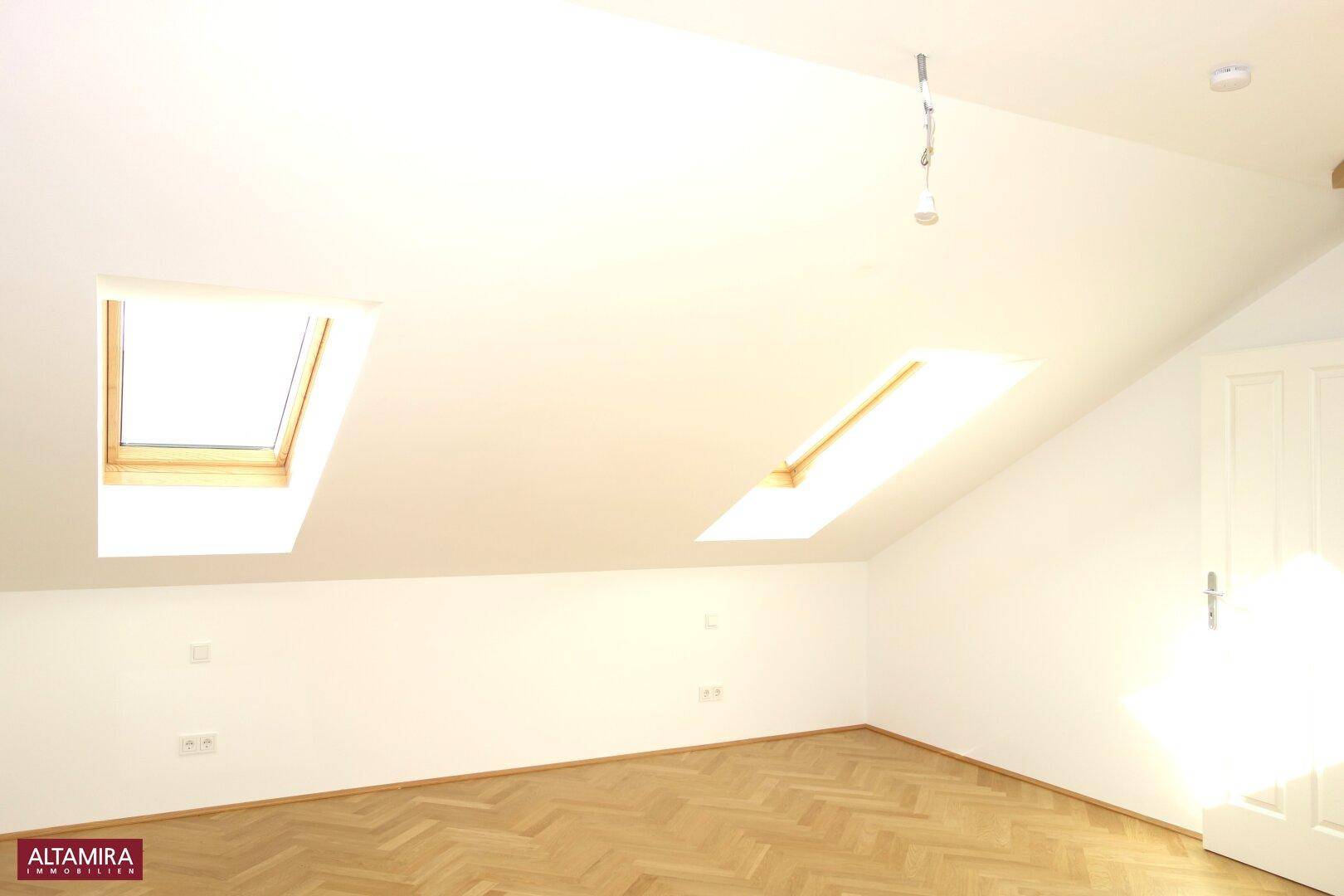 Helles Dachgeschosszimmer mit zwei Dachfenstern, schrägen Wänden und elegantem Fischgrätparkettboden.