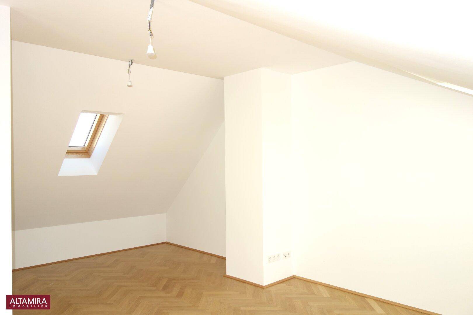 Dachgeschossraum mit schrägen Wänden, Fischgrätparkett und einem Dachfenster für natürliches Licht.