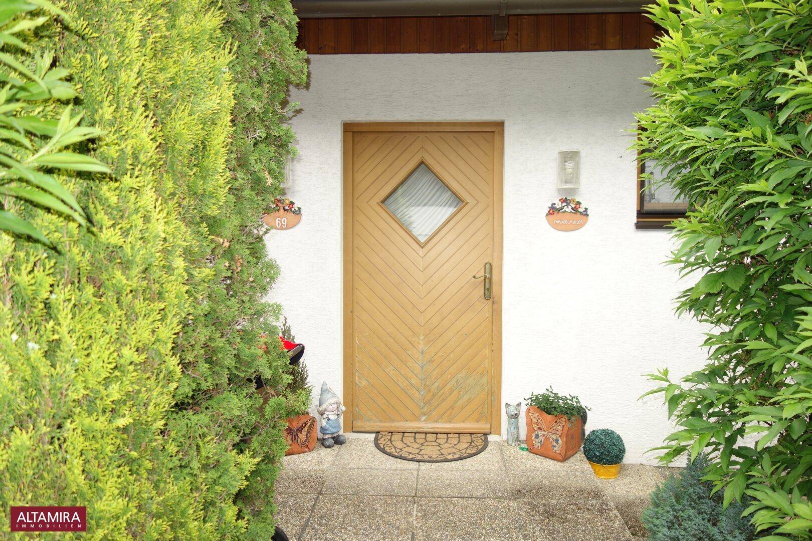 Hauseingang mit Holztür, umgeben von grünen Hecken und kleinen Gartenzwergen.