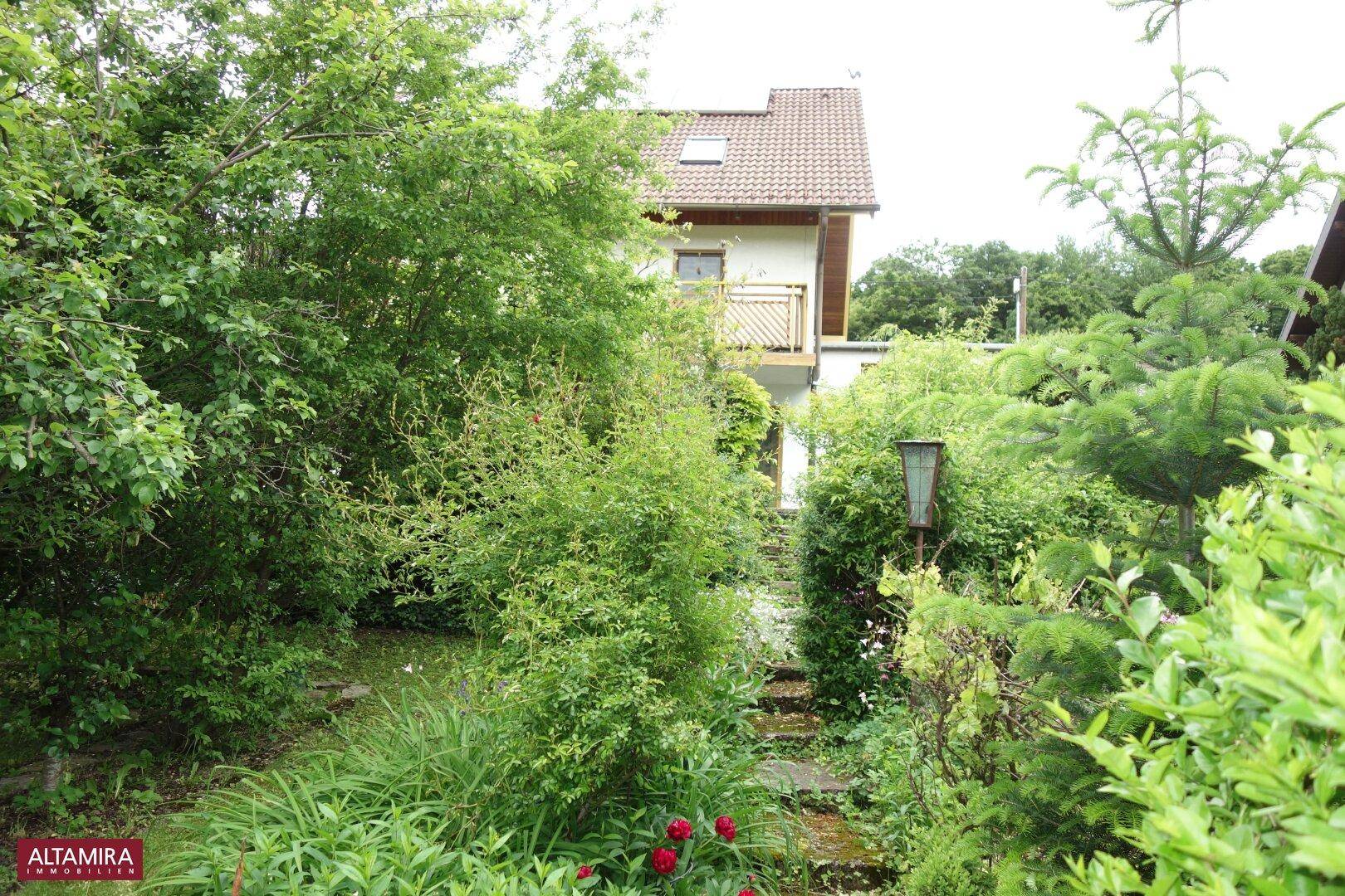 Verwunschener Gartenweg führt durch üppiges Grün zu einem Haus mit Balkon.