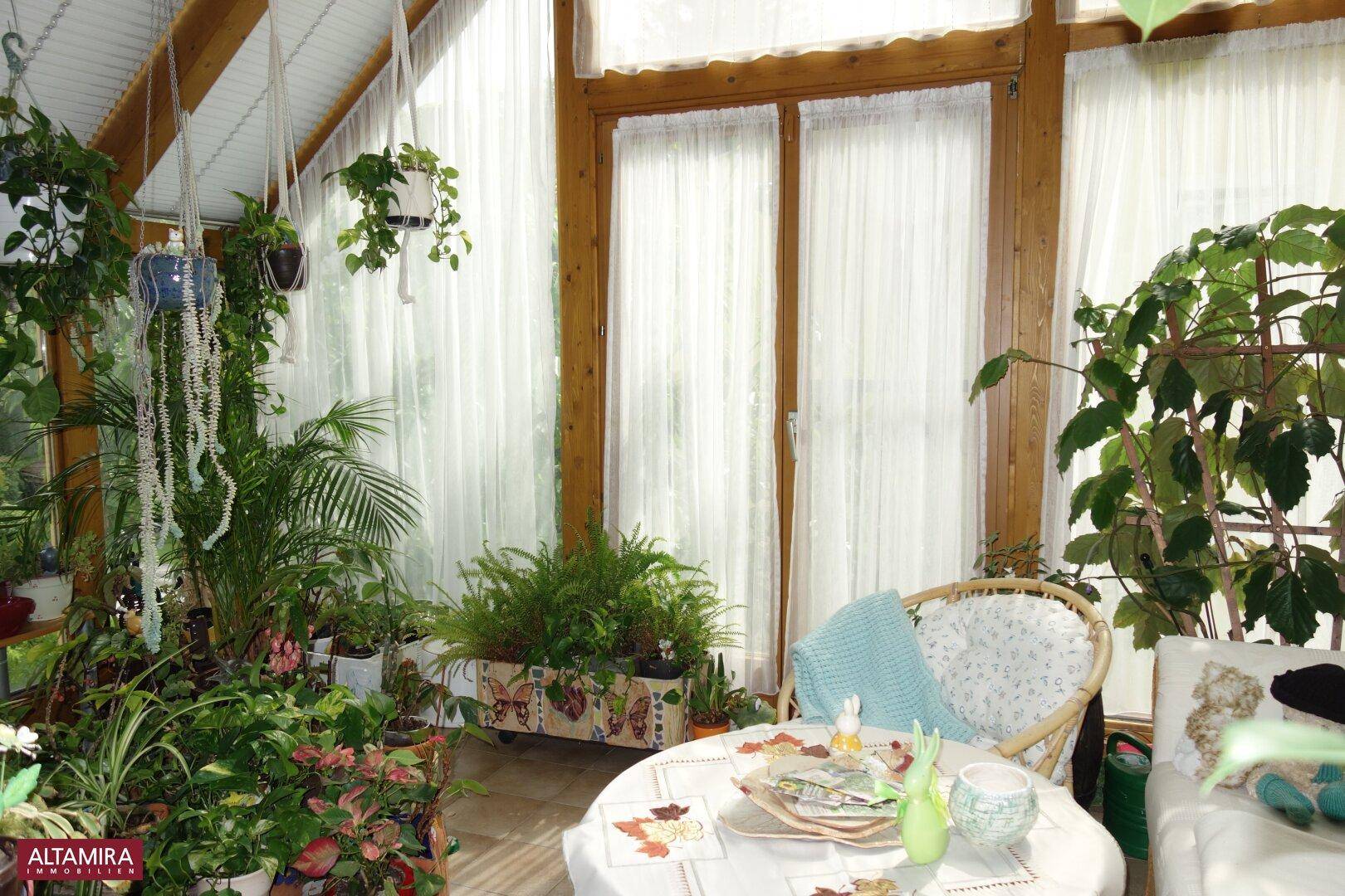 Heller Wintergarten mit Rattanmöbeln, üppiger Bepflanzung und Blick ins Grüne.