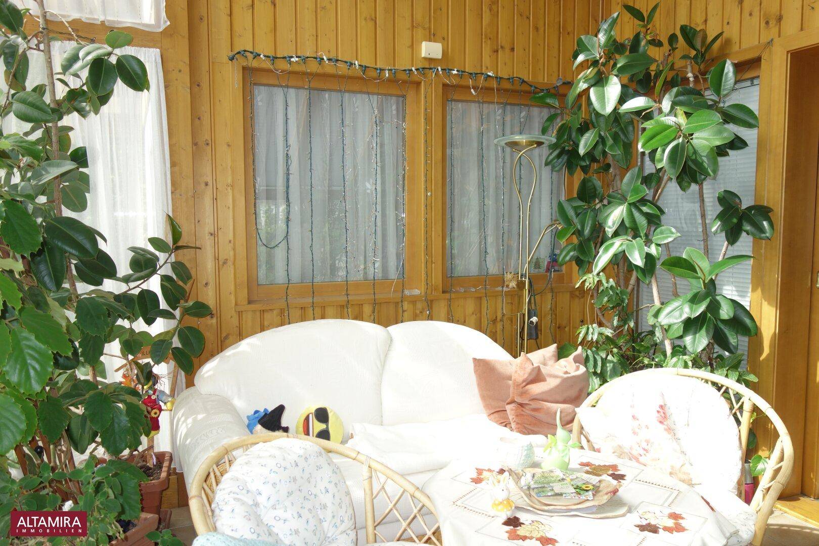 Gemütliche Sitzecke im Wintergarten mit Rattanmöbeln, vielen Pflanzen und Holzverkleidung.