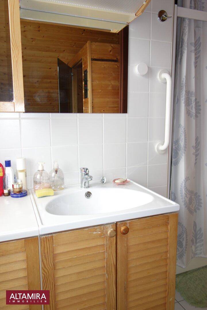 Badezimmer mit Waschtisch, Holzschränken, Spiegel und Haltegriff an der gefliesten Wand.