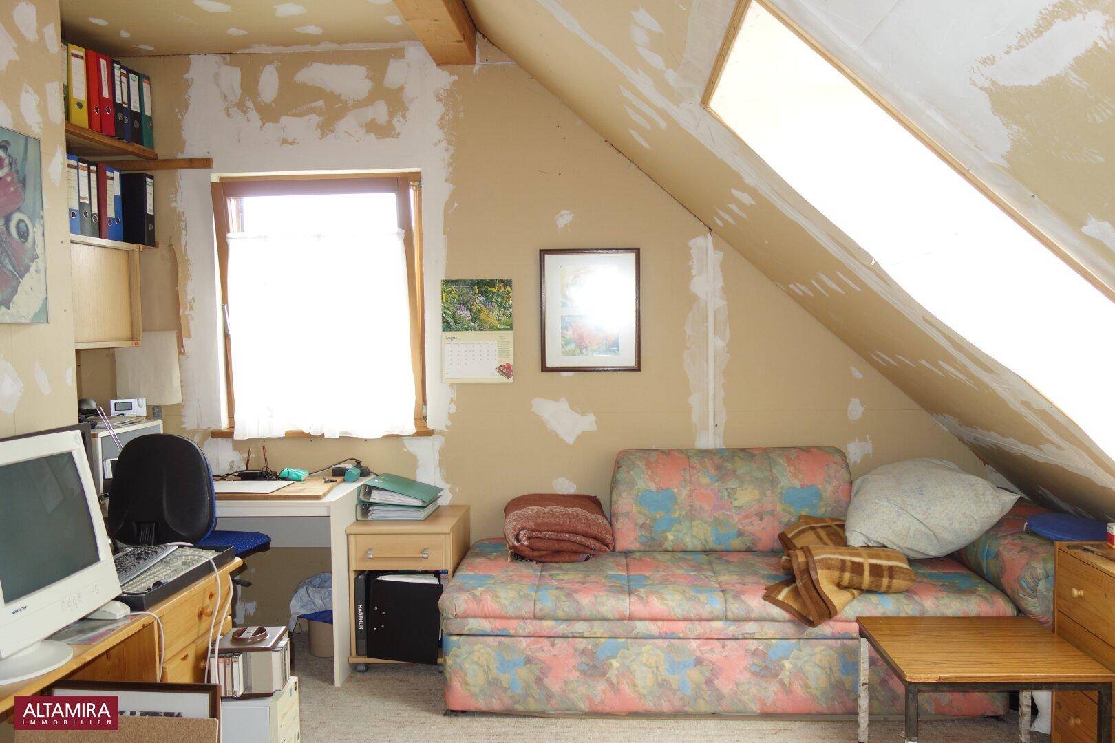 Dachzimmer mit schrägen Wänden, Dachfenster, Schreibtisch und Schlafsofa, renovierungsbedürftig.