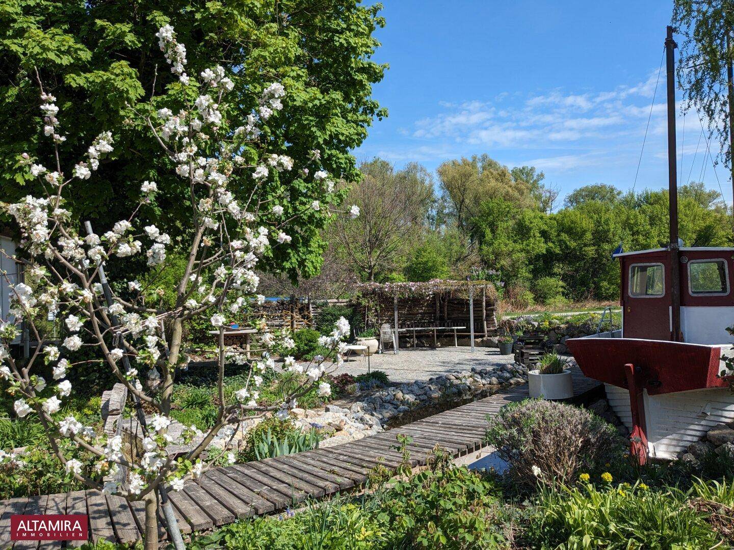 Blühender Garten mit Holzsteg über einem kleinen Teich und einem dekorativen Boot im Hintergrund.