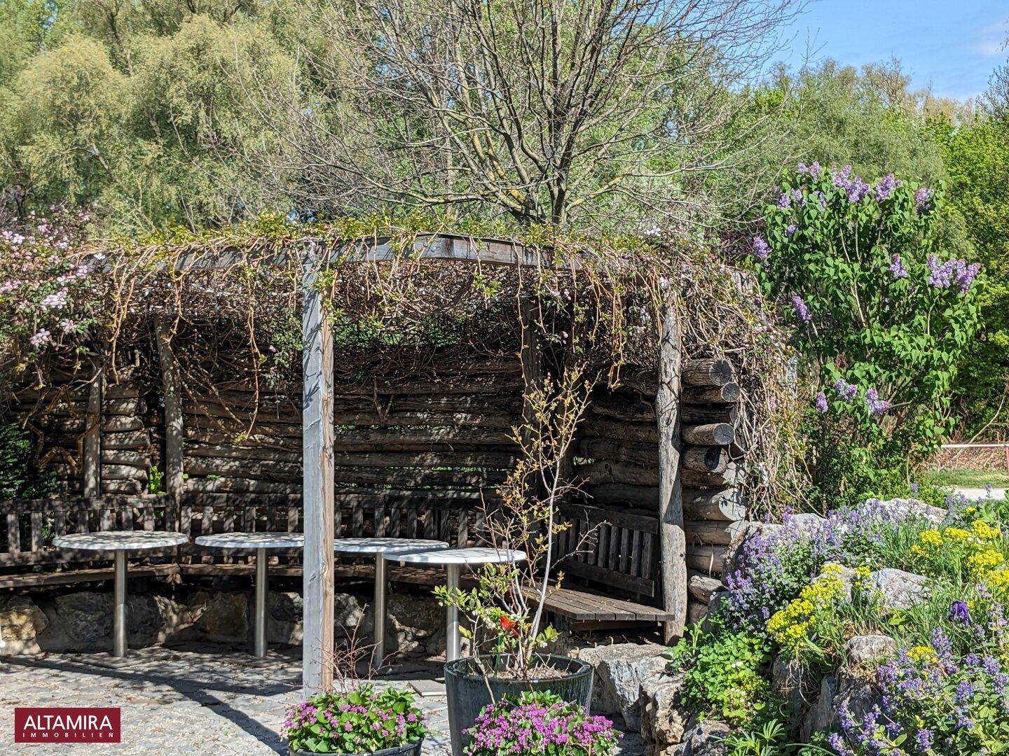 Gemütlicher Sitzbereich im Garten unter einer bewachsenen Holzpergola, umgeben von Blumen.
