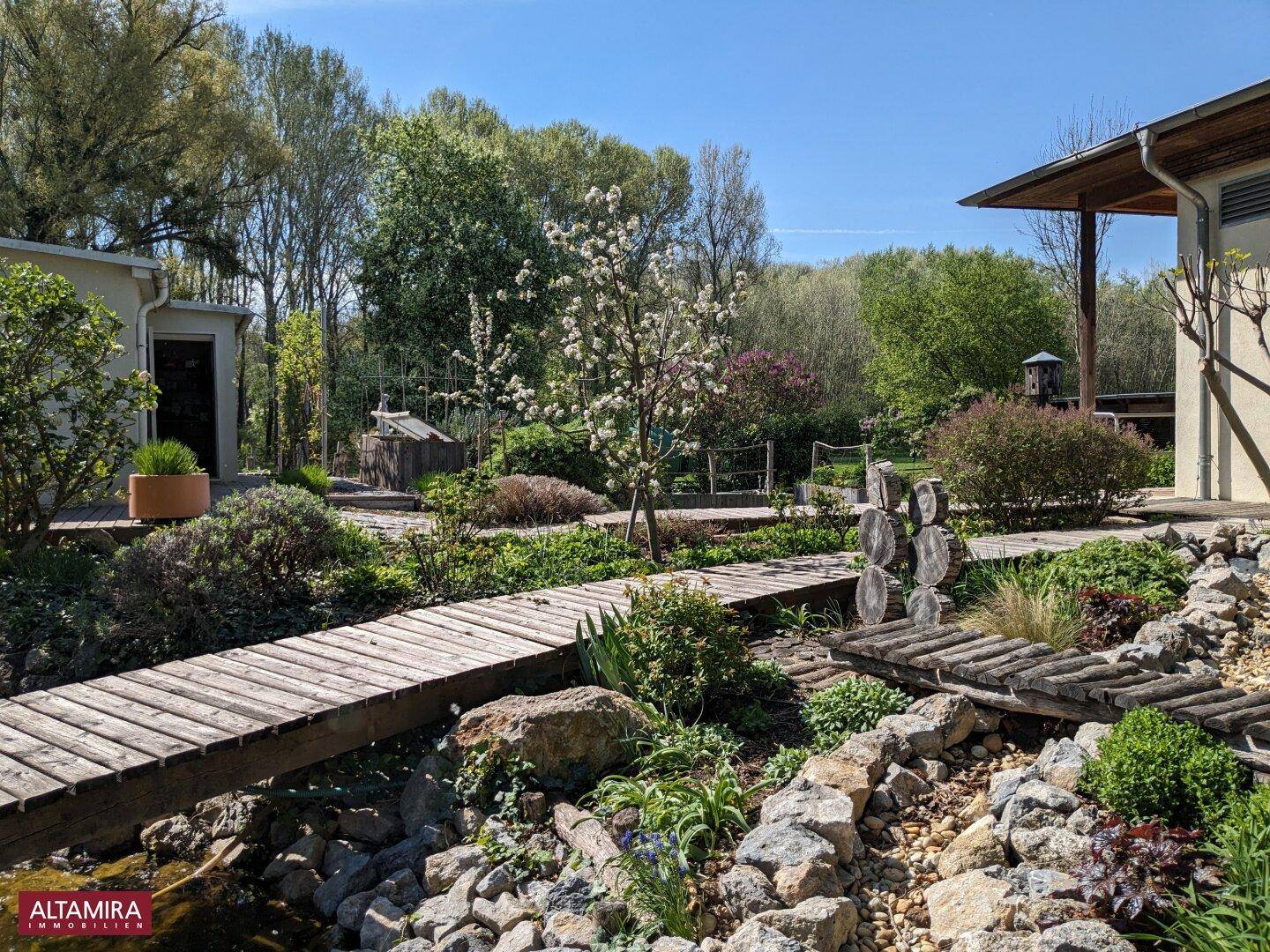Gepflegter Garten mit Holzsteg über einem Bachlauf, üppiger Bepflanzung und dem Gebäude im Hintergrund.