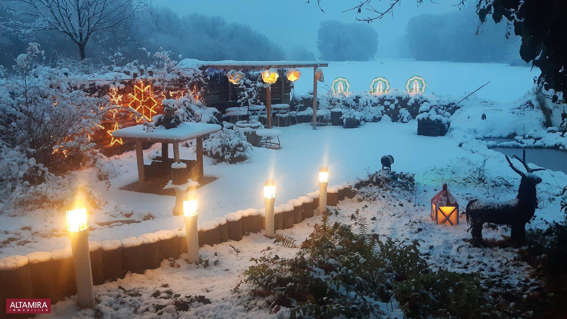 Winterlicher Garten, bedeckt mit Schnee und festlicher Weihnachtsbeleuchtung in der Dämmerung.