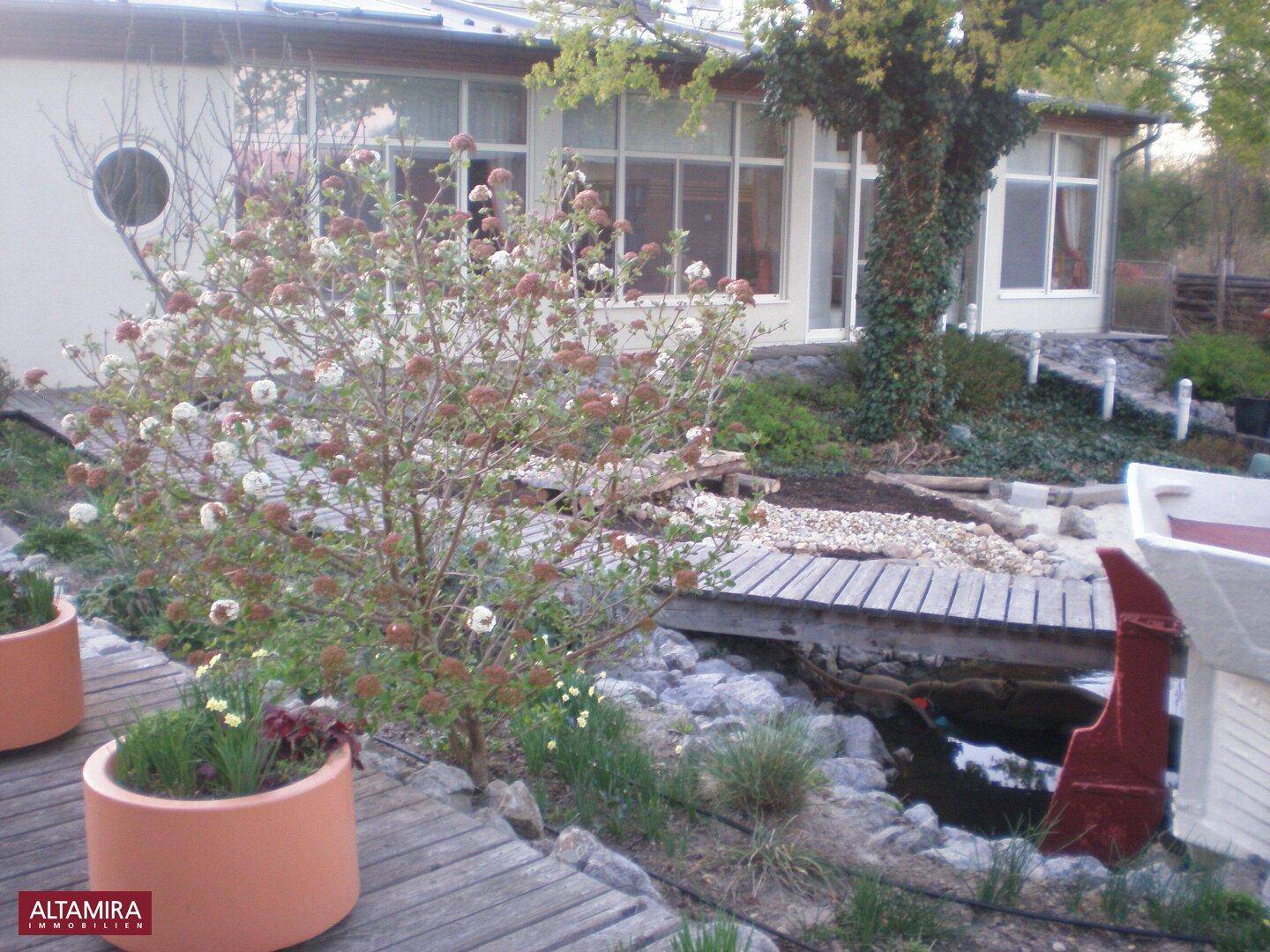 Gartenbereich mit Holzsteg über einen kleinen Teich und Blick auf die Gebäudeseite mit großen Fenstern.