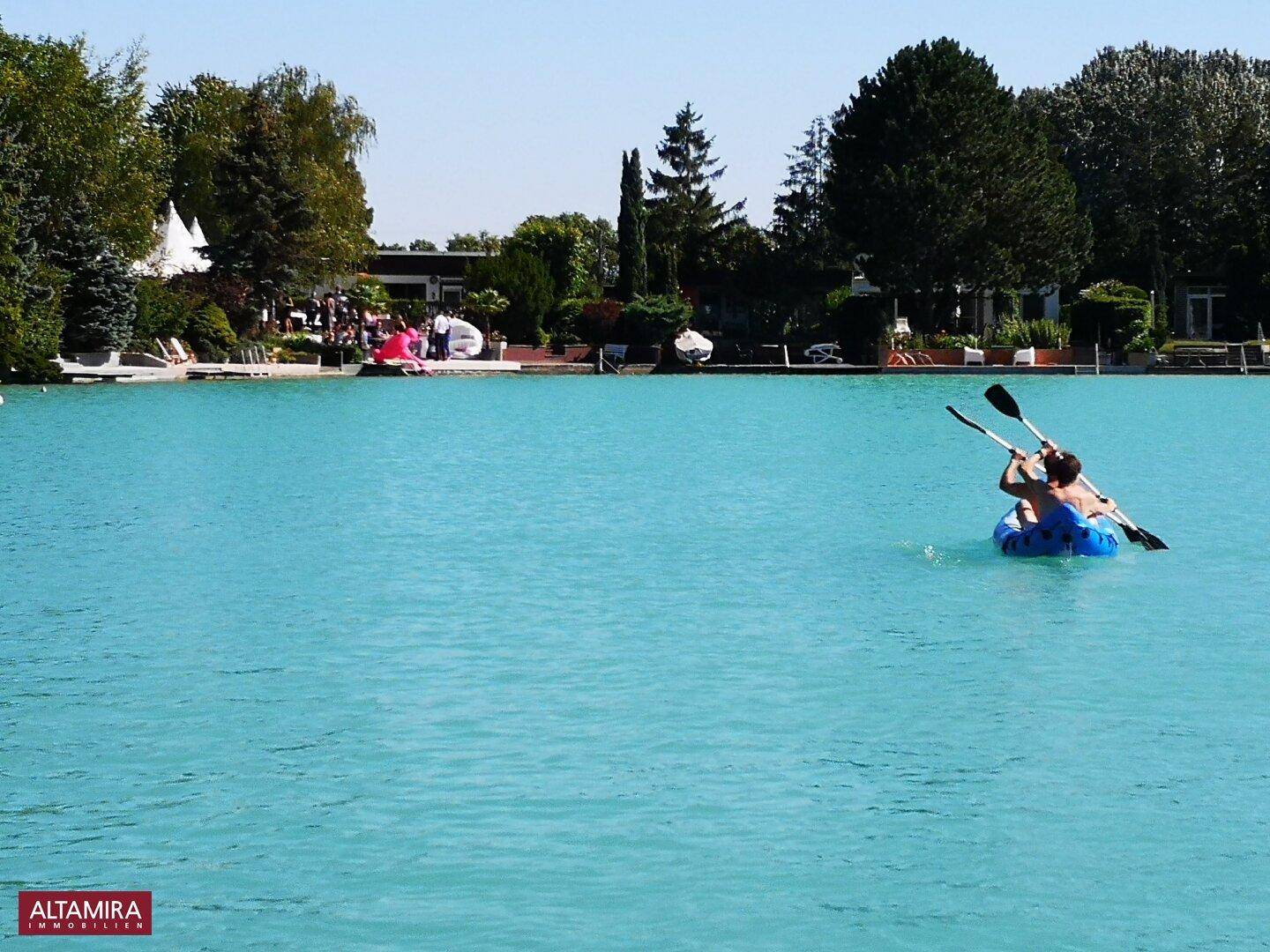 Blauer See mit Menschen beim Kajakfahren und einer grünen Uferlandschaft im Hintergrund.
