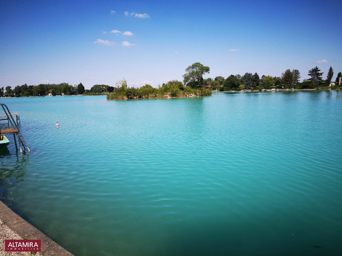 Weitläufiger blauer See mit einer kleinen Insel und bewaldetem Ufer unter klarem Himmel.