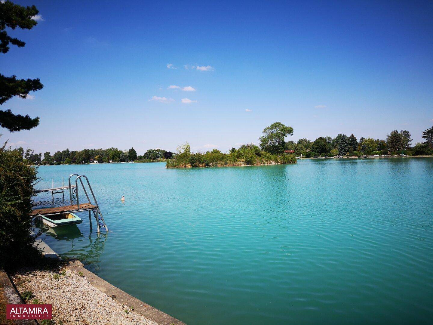 Malerischer blauer See mit Badeleiter, kleiner Insel und grüner Uferlandschaft.