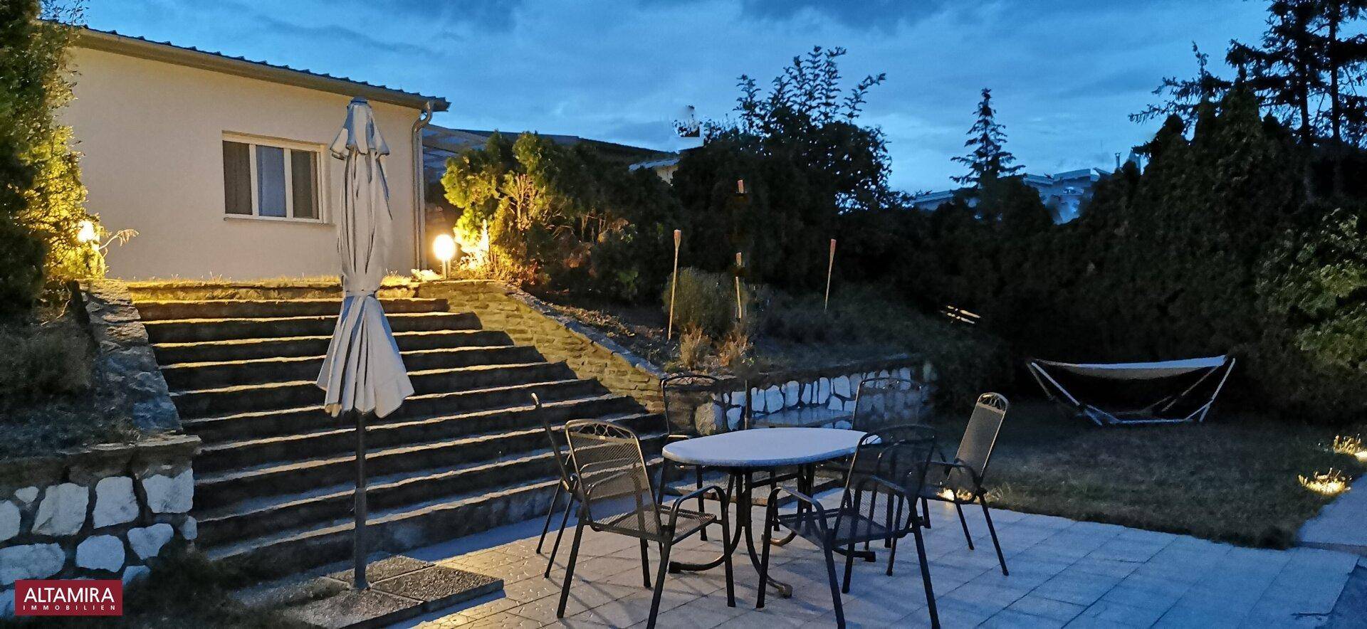 Beleuchtete Terrasse am Abend mit Gartenmöbeln und Treppen, die zum Haus führen.