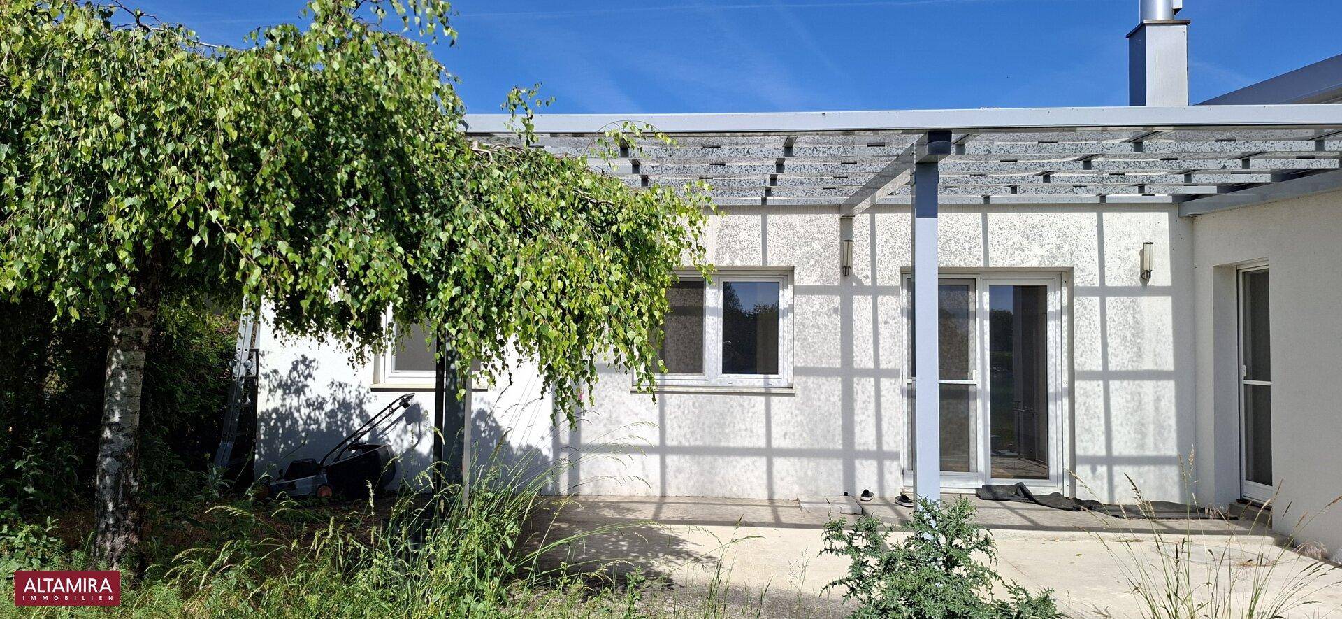 Seitenansicht des Hauses mit weißer Fassade, Terrassentüren und einer Pergola im Gartenbereich.