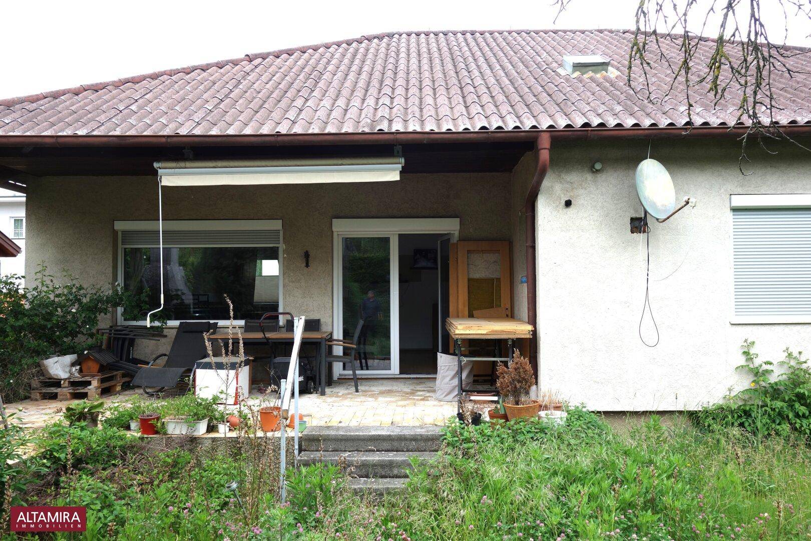 Überdachte Terrasse mit Gartenmöbeln und Blick auf den verwilderten Gartenbereich.