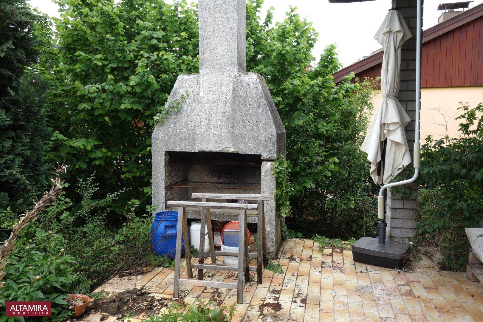 Gemauerter Grillplatz im Garten auf gepflasterter Fläche, umgeben von üppigem Grün.