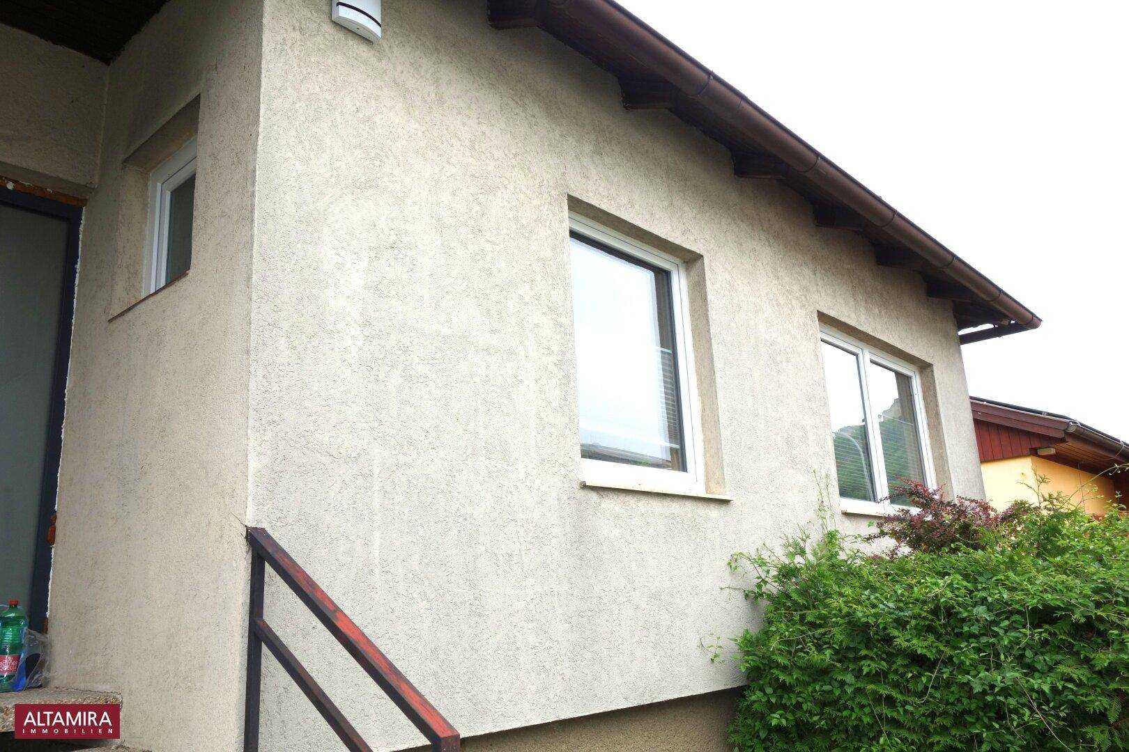 Eckansicht des Hauses mit Fenstern und einer Außentreppe, umgeben von Sträuchern.