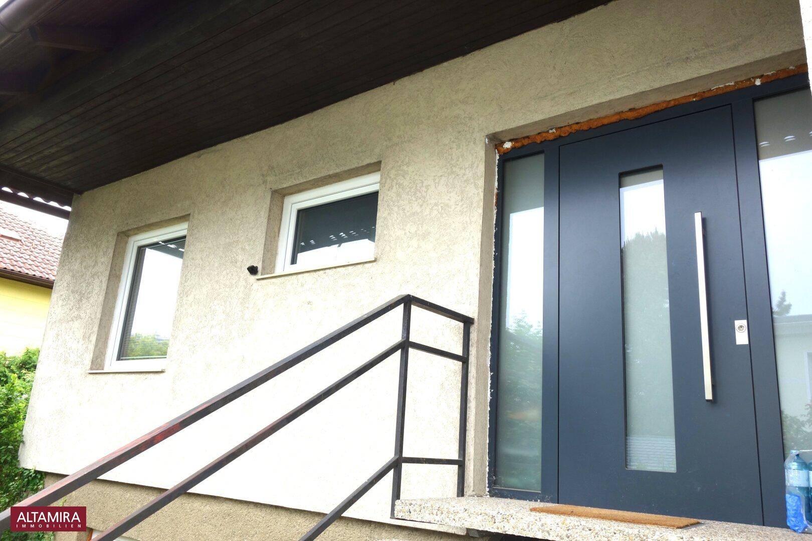 Moderner Hauseingang mit dunkler Tür und seitlichen Glaselementen, zugänglich über eine Treppe.