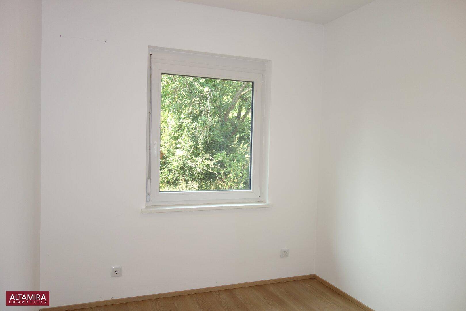 Helles Schlafzimmer mit Laminatboden und Fenster, das einen ruhigen Blick ins Grüne bietet.