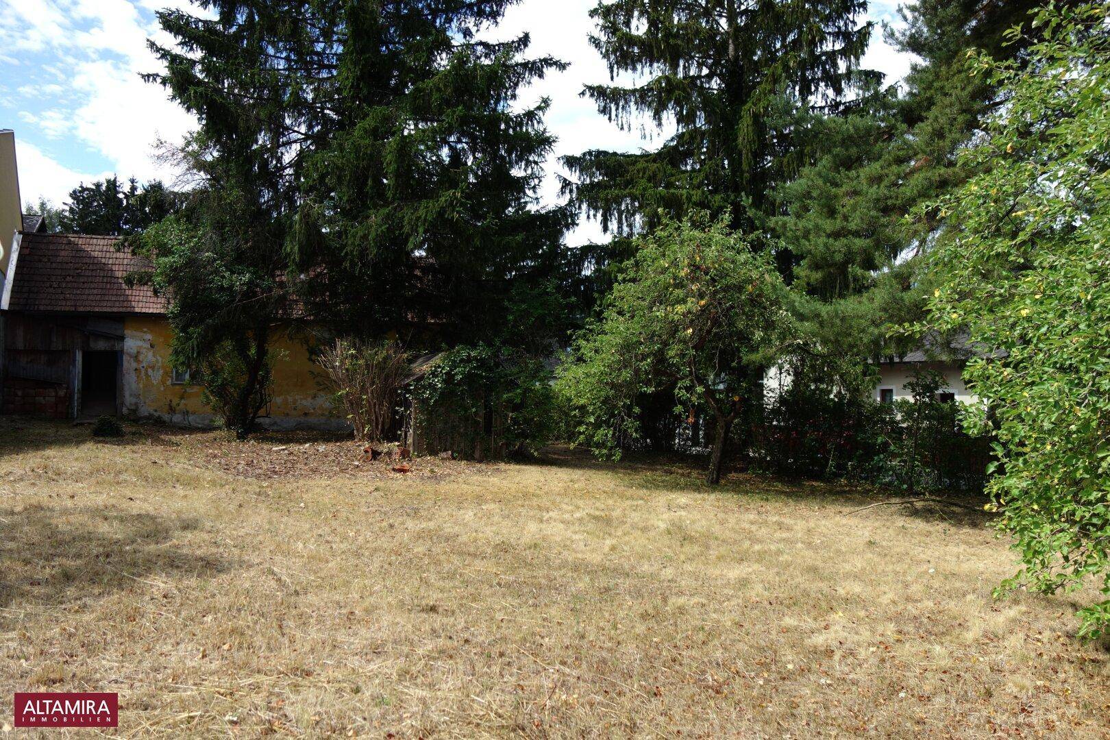 Großer Garten mit alten Bäumen und einem verputzten gelben Gebäude im Hintergrund.