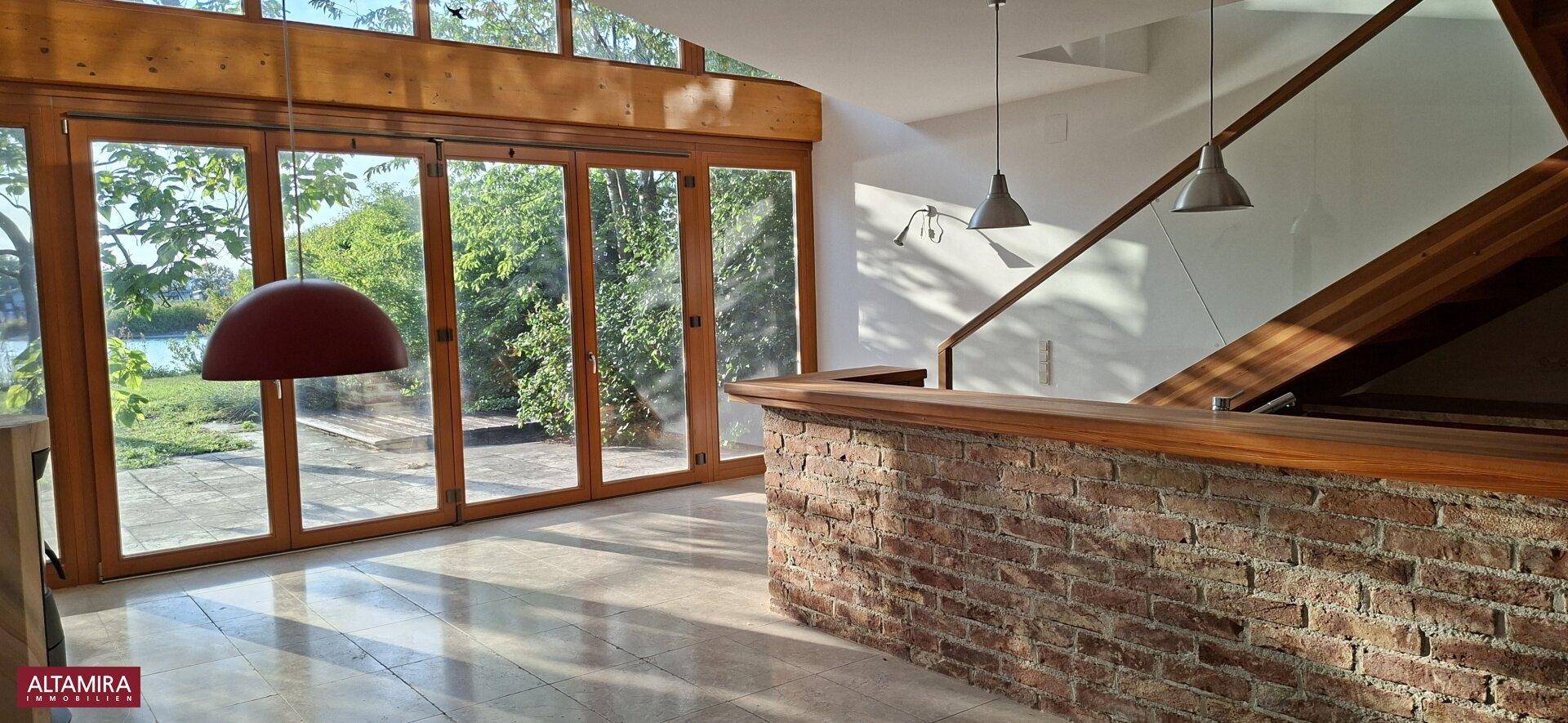 Heller Wohnbereich mit großen Glasfronten, Seeblick, einer Ziegelbar und Holztreppe.