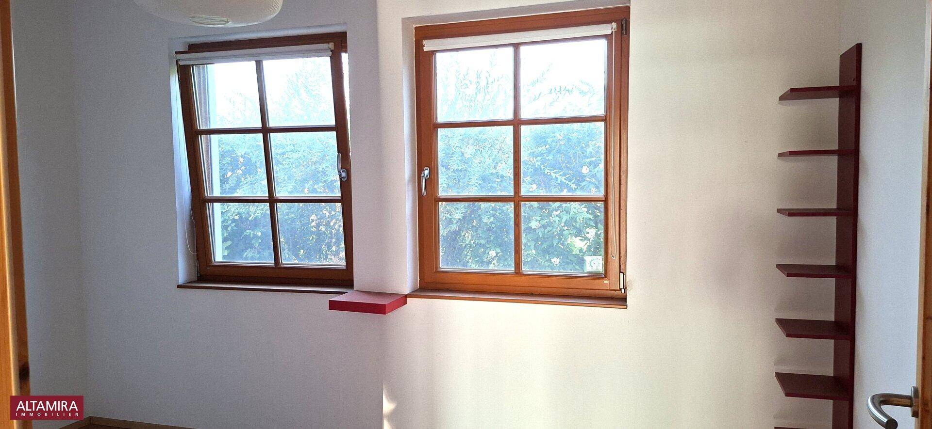 Heller Raum mit zwei Fenstern und einer schlichten Holzablage an der Wand.