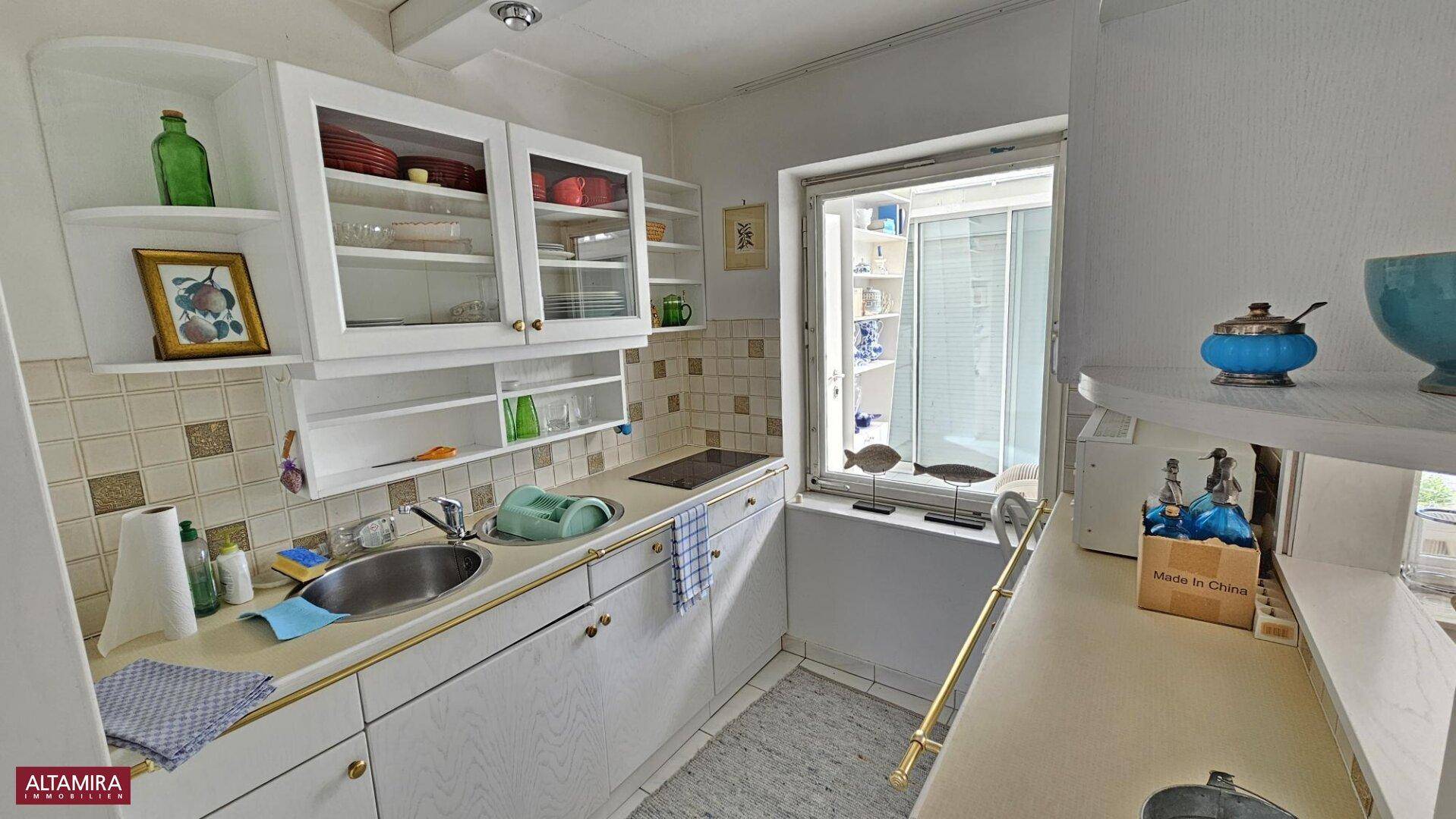 Kleine Küche mit weißen Schränken, Fliesenrückwand und einem Fenster mit Blick nach draußen.