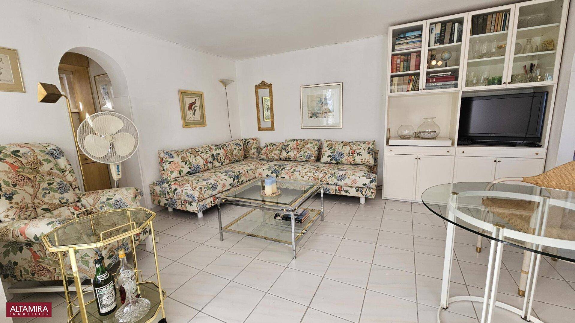 Gemütliches Wohnzimmer mit floral gemustertem Sofa, Fliesenboden und einem Bücherregal.