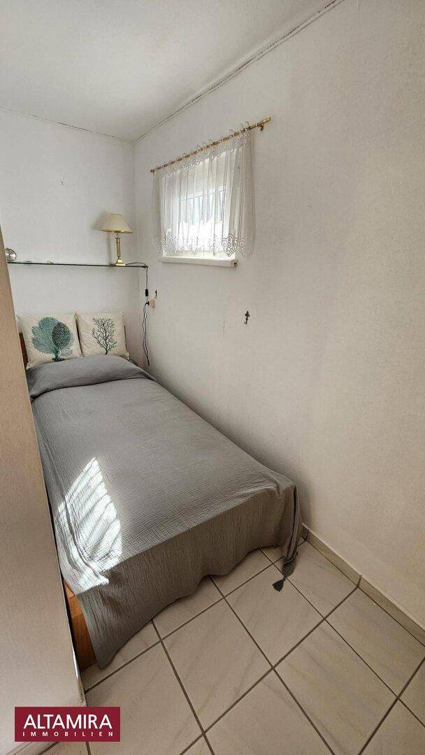 Kleines Schlafzimmer mit Einzelbett, Fliesenboden und einem Fenster mit Vorhang.