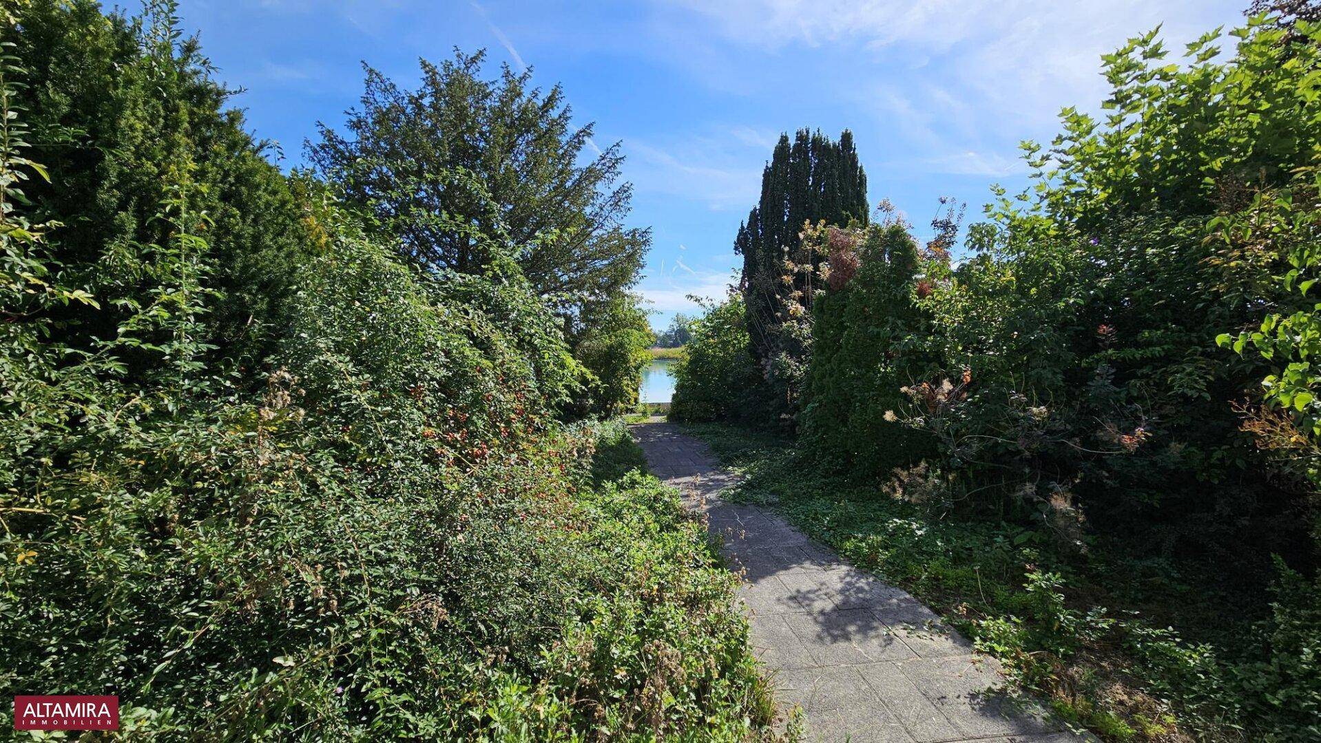 Idyllischer Gartenweg umgeben von Bäumen und Sträuchern mit Blick auf den See.