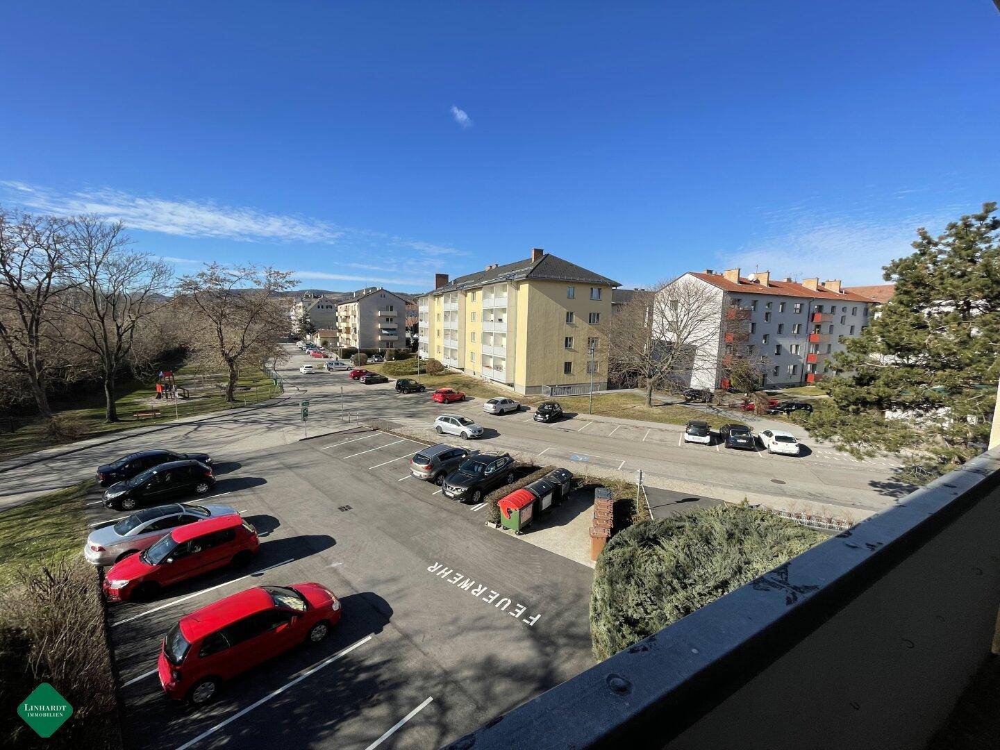Blick von einem Balkon auf die Straße mit parkenden Autos und umliegenden Wohngebäuden unter blauem Himmel.