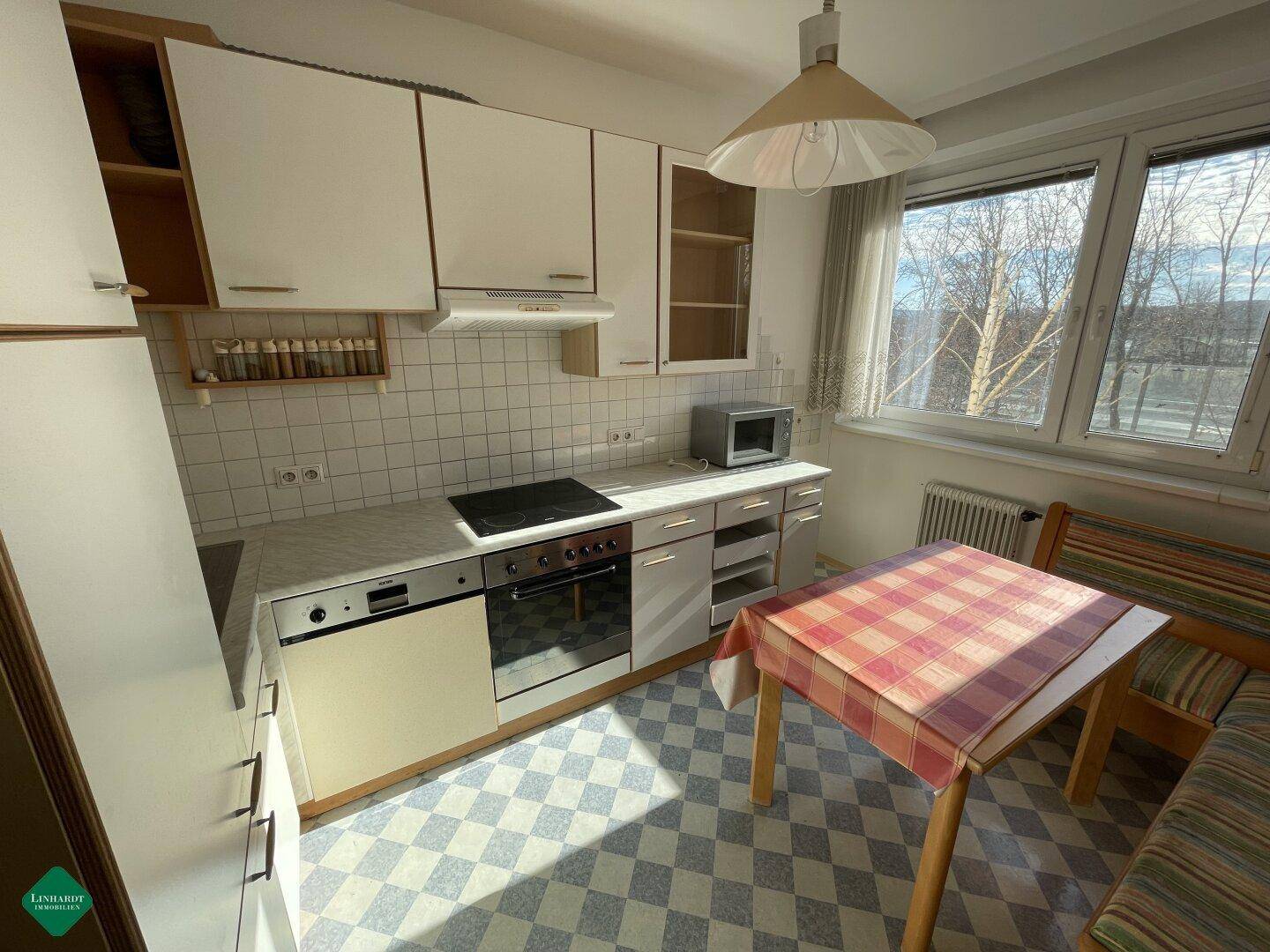 Helle Küche mit weißen Schränken, kariertem Boden und kleinem Essbereich am Fenster.