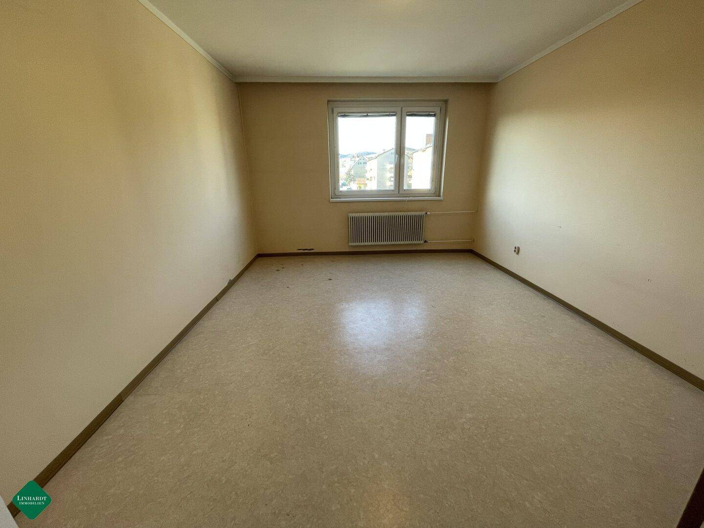 Leerer Raum mit beigen Wänden, einem Fenster und hellem Bodenbelag.
