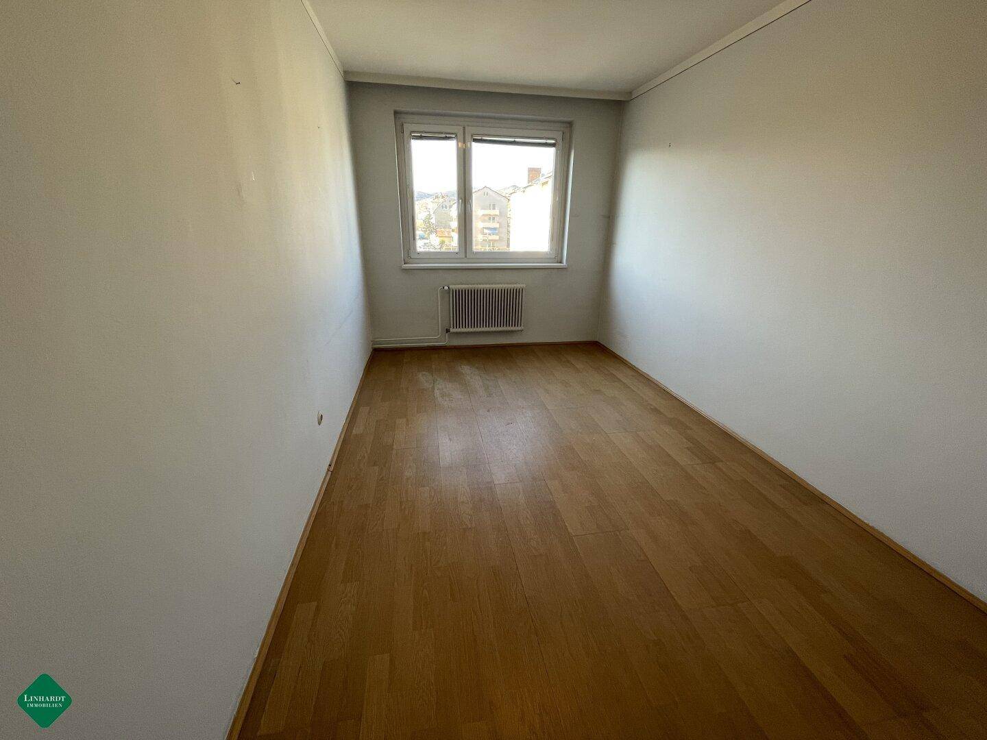 Leerer Raum mit hellgrauen Wänden, einem Fenster und Holzfußboden.