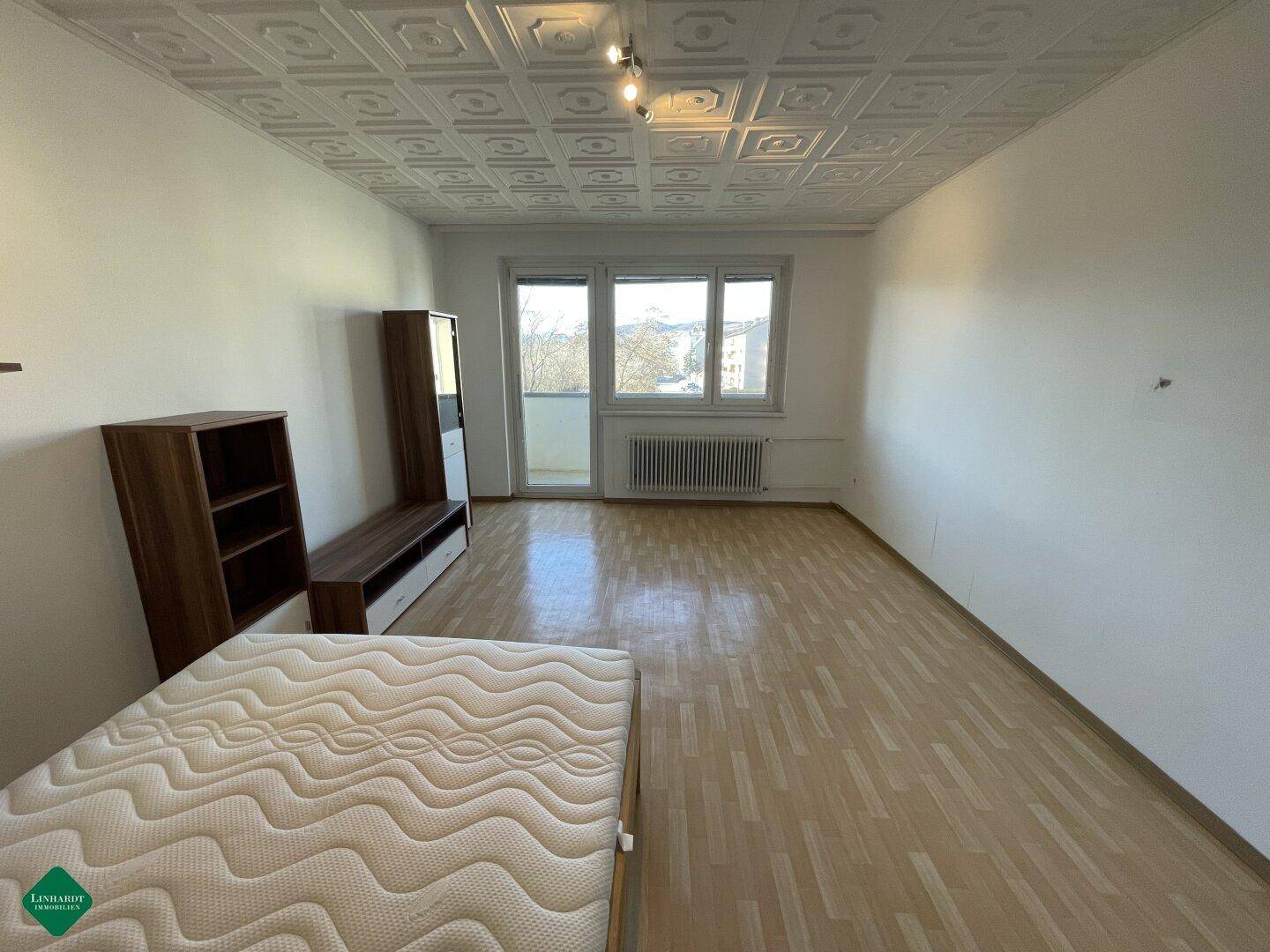 Schlafzimmer mit Matratze auf dem Boden, Holzregalen und Zugang zu einem kleinen Balkon.