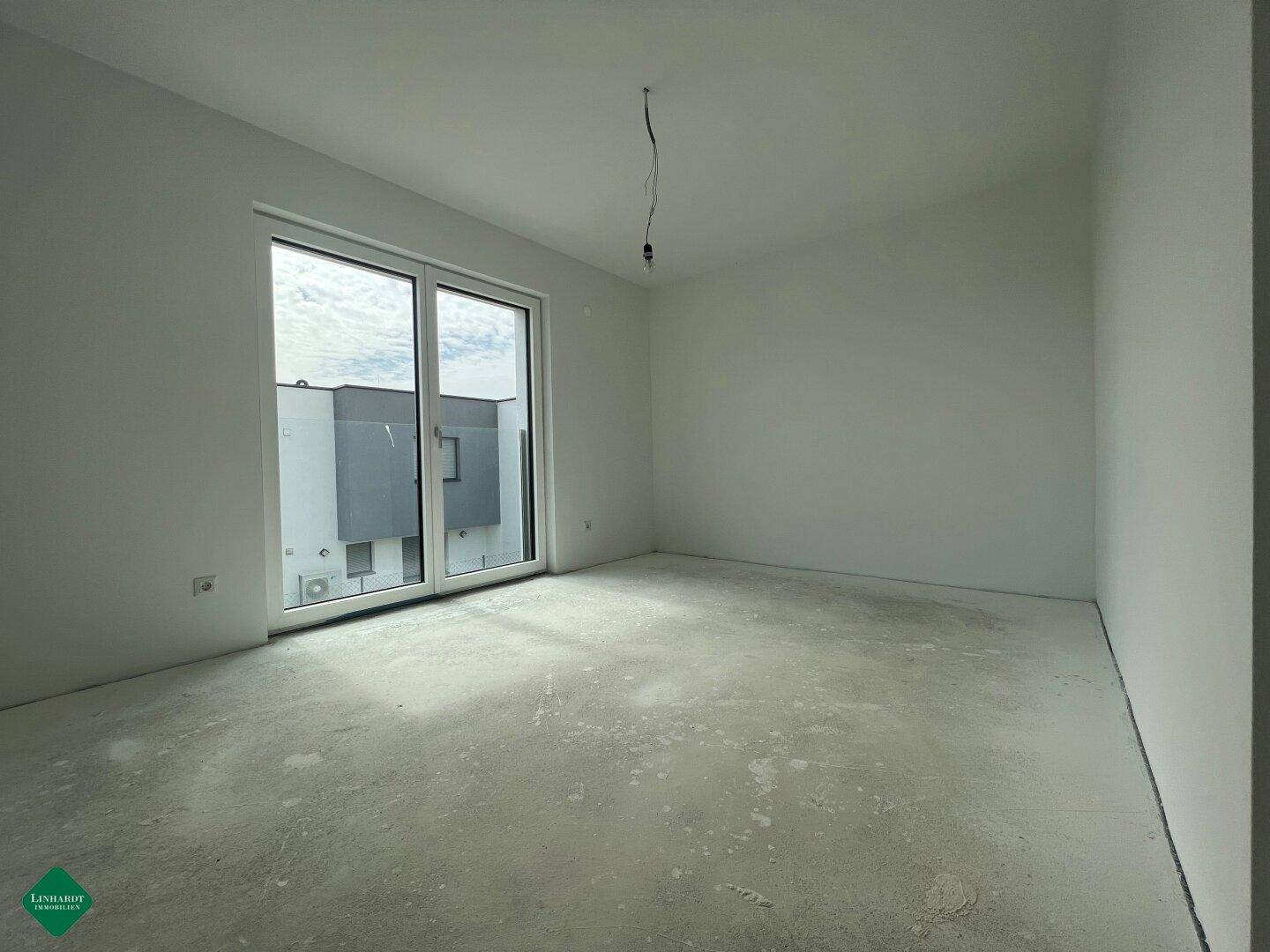 Helles Schlafzimmer im Rohbau mit großem Fenster und Blick auf ein angrenzendes Gebäude.