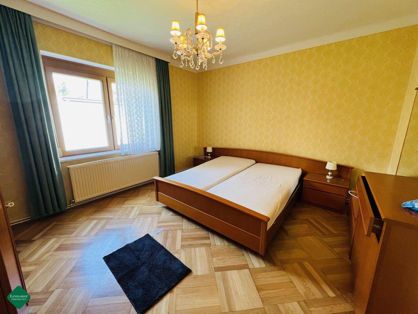 Schlafzimmer mit Doppelbett, gemusterten gelben Tapeten und Parkettboden.