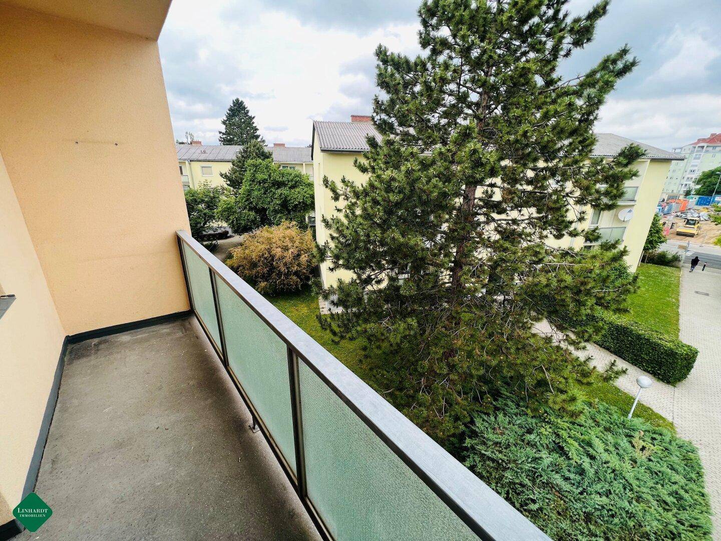 Balkon mit Glasbrüstung und Blick ins Grüne, umgeben von Bäumen und Wohngebäuden.