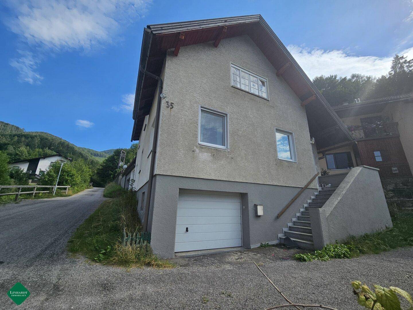 Graues Wohnhaus mit Garage im Erdgeschoss und Außentreppe in ländlicher Umgebung.